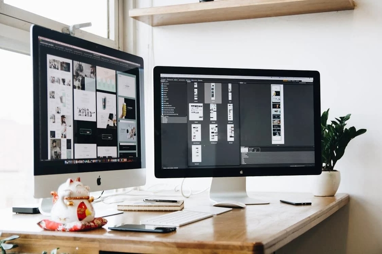 Dois monitores em uma mesa de trabalho exibindo um software de design com múltiplos wireframes e componentes, ilustrando o processo de prototipagem